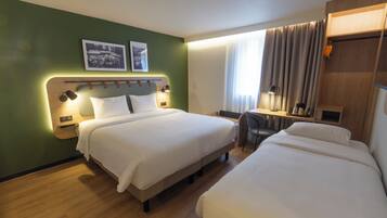 A hotel room with two beds, a desk, and a chair. There are two framed pictures on the wall, a window with curtains, and a wooden cabinet.