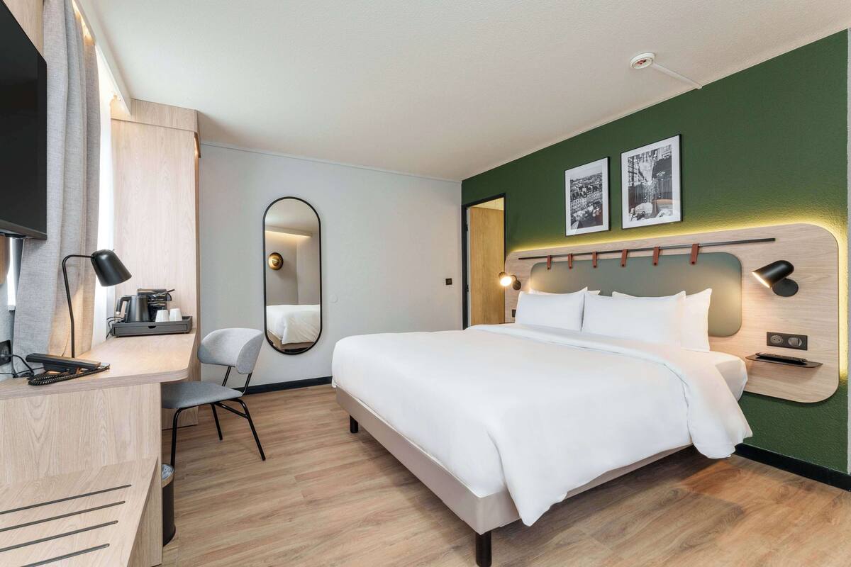 Ein modernes Hotelzimmer mit einem großen Bett, einem Schreibtisch mit Stuhl, einem Spiegel und einem Fernseher.