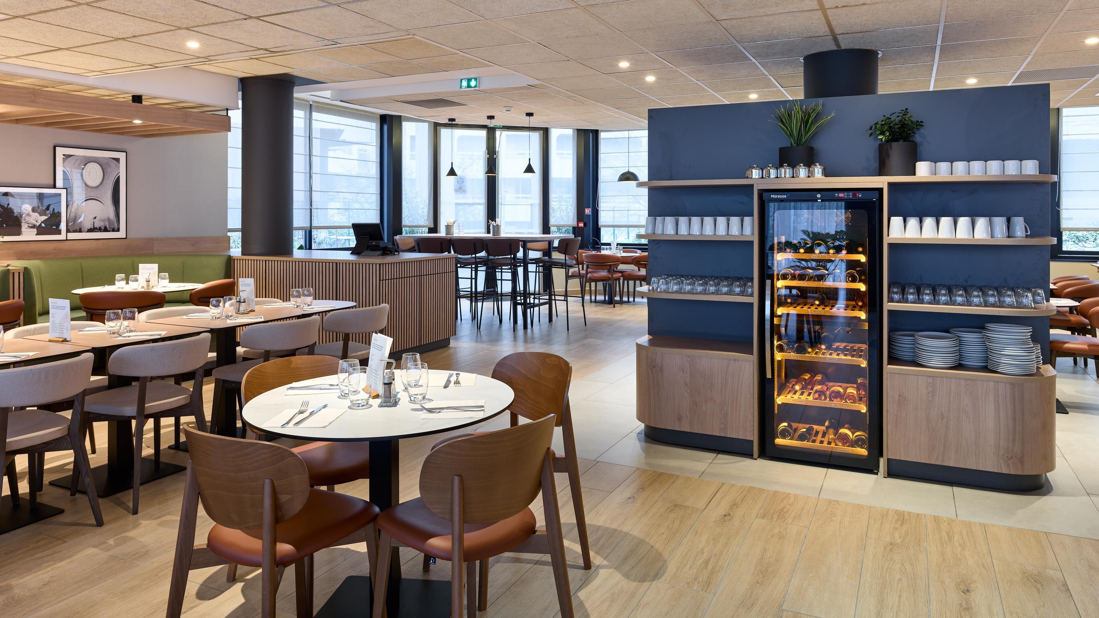 Restaurante moderno com mesas redondas, cadeiras de madeira e uma ilha central. Há um expositor com copos e pratos.
