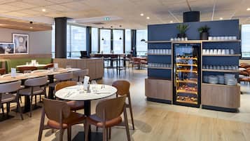 Restaurante moderno com mesas redondas, cadeiras de madeira e uma ilha central. Há um expositor com copos e pratos.