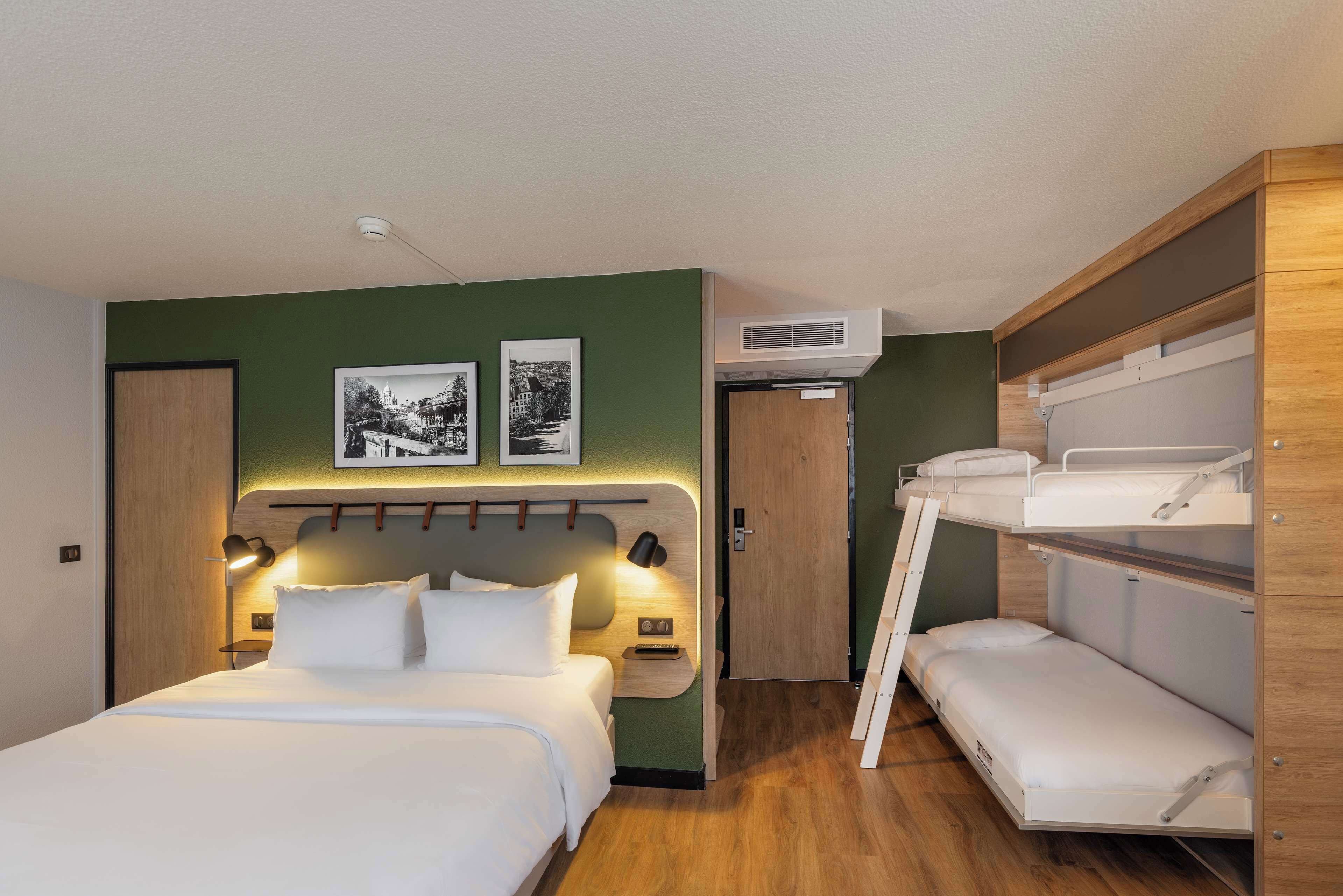 A modern hotel room with a large bed, a bunk bed, a desk with a lamp, and two framed pictures on the wall.