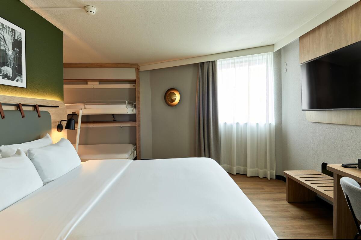 Ein Hotelzimmer mit einem großen Bett, einem Schreibtisch, einem Flachbildfernseher, Stockbetten und einer Wanduhr.
