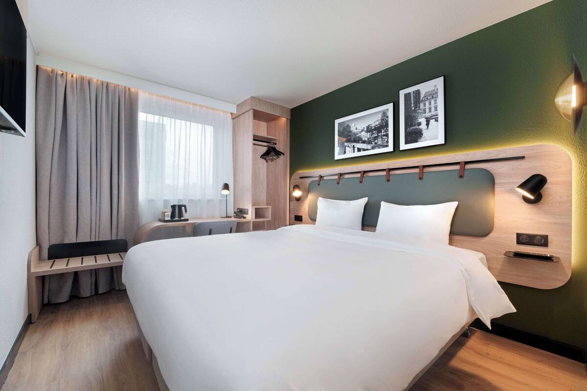 Ein modernes Hotelzimmer mit einem großen Bett, einem Schreibtisch, einem Sessel, einem Fenster mit Vorhängen und einem an der Wand befestigten Fernseher.