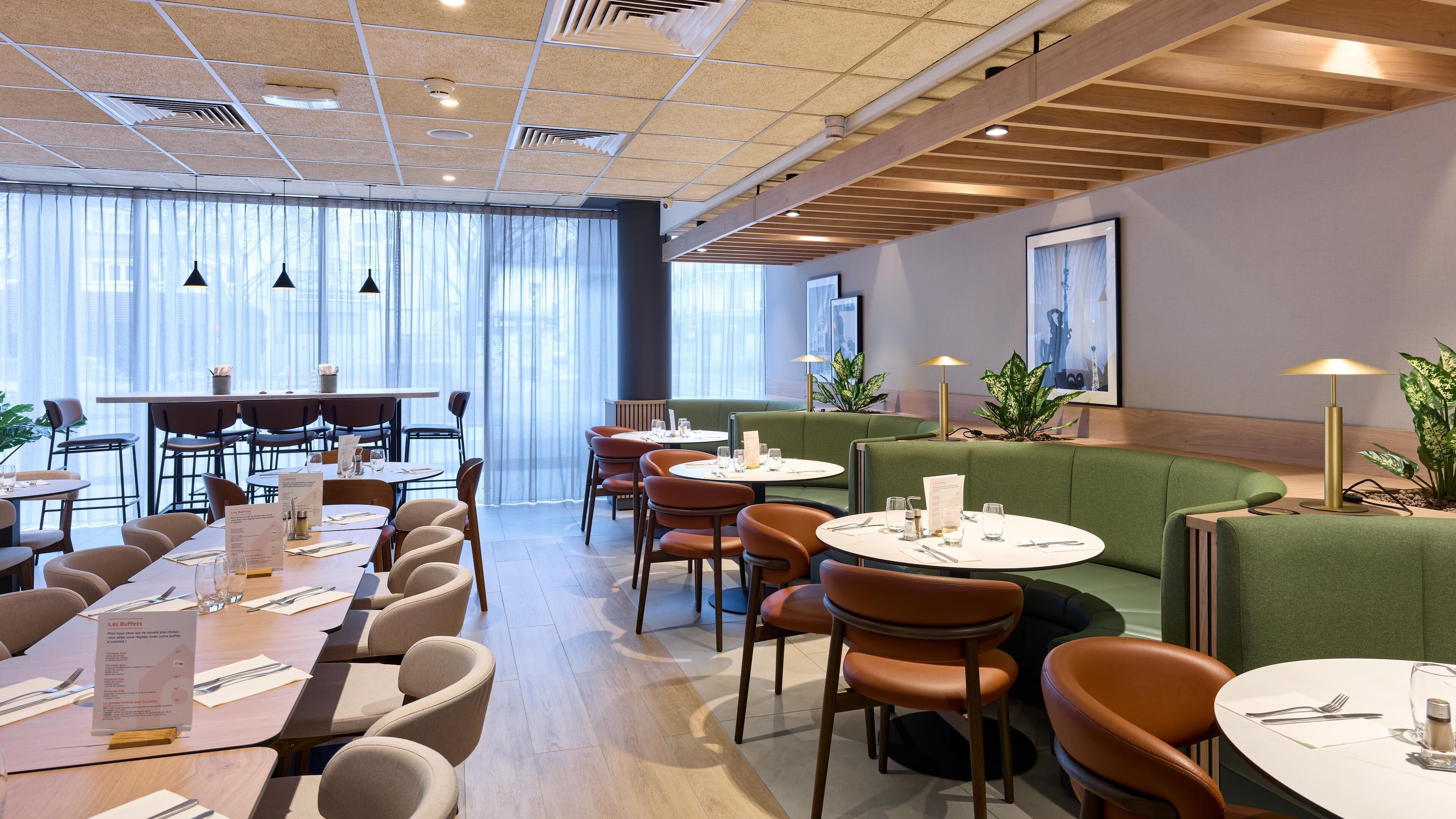 Un restaurant moderne avec des tables rondes et des banquettes, de grandes fenêtres et des plantes d’intérieur.