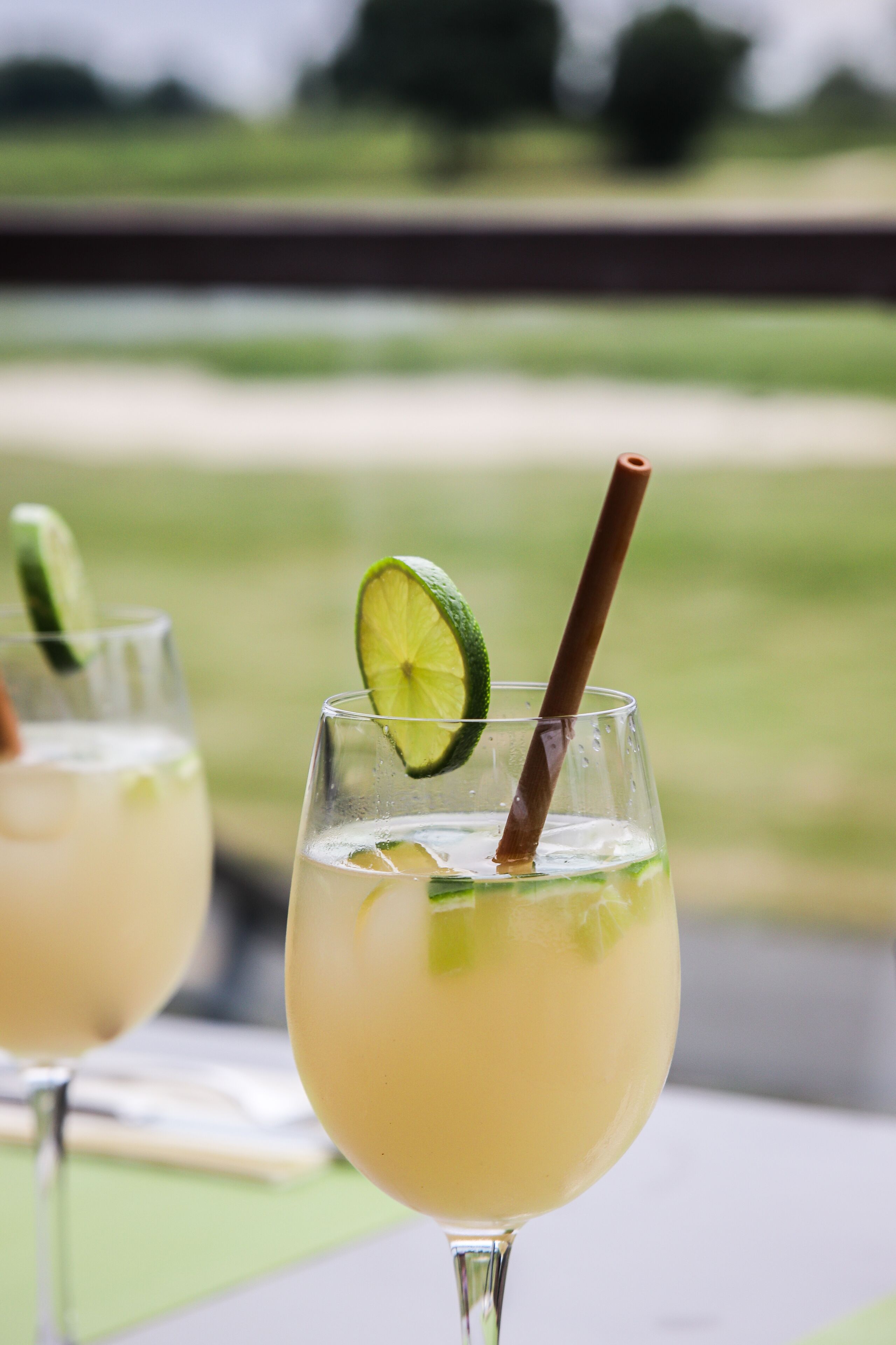 Deux verres contenant une boisson claire, agrémentée de rondelles de citron vert et de pailles, sont placés sur un fond extérieur flou.