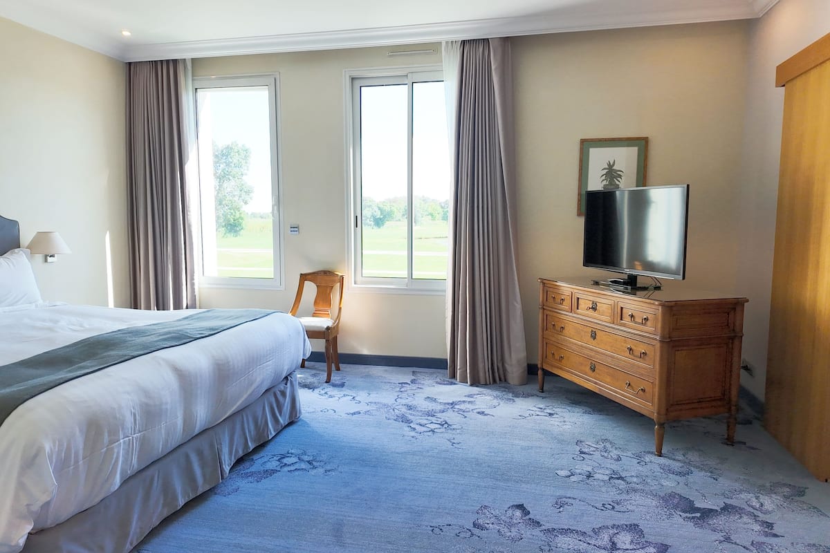 Une chambre d’hôtel avec un grand lit, une commode en bois avec un téléviseur, deux fenêtres avec des rideaux et une vue sur l’extérieur.