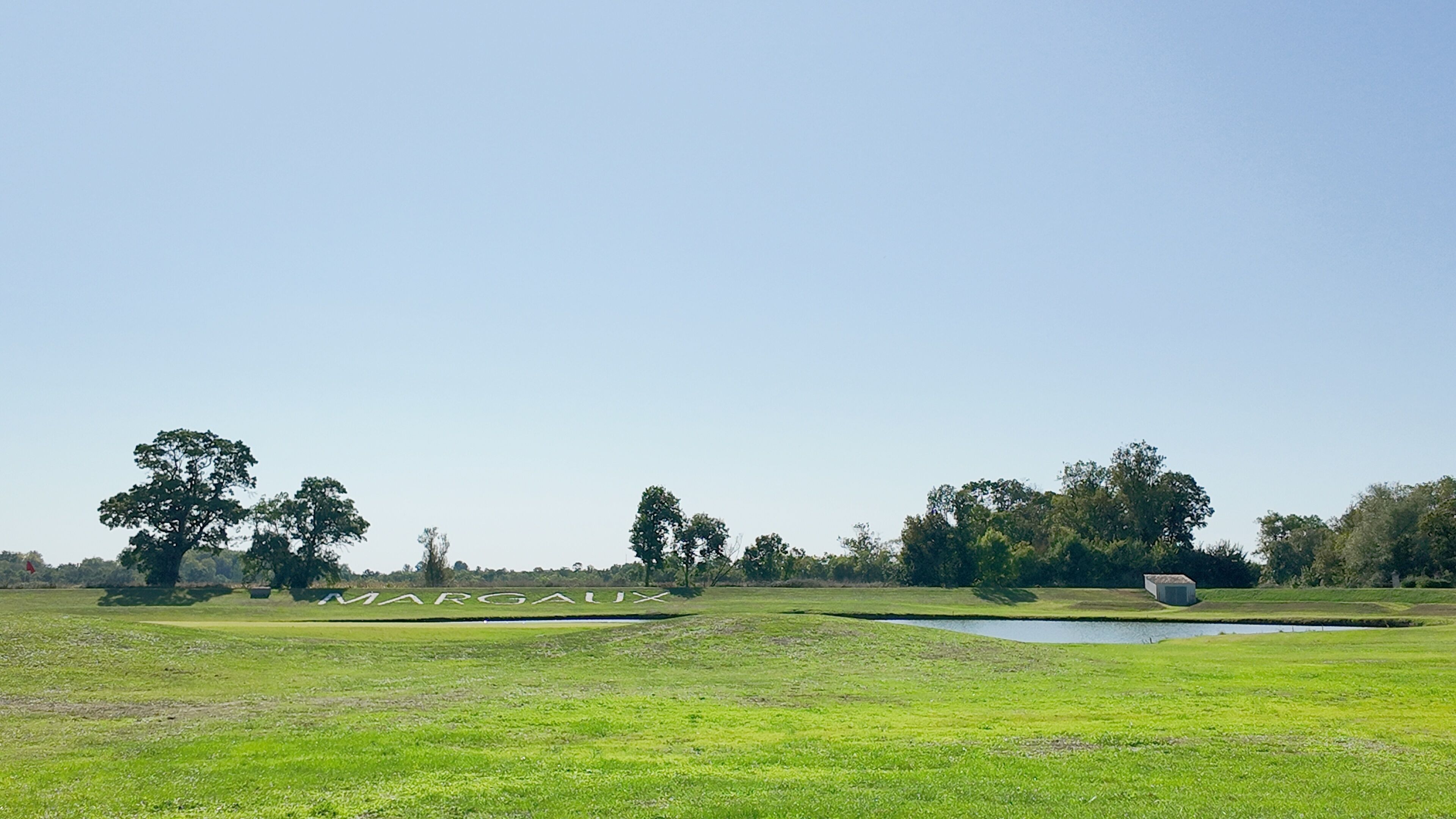 Ein Golfplatz mit Teich, Bäumen und dem Schriftzug "MARGAUX" im Gras.