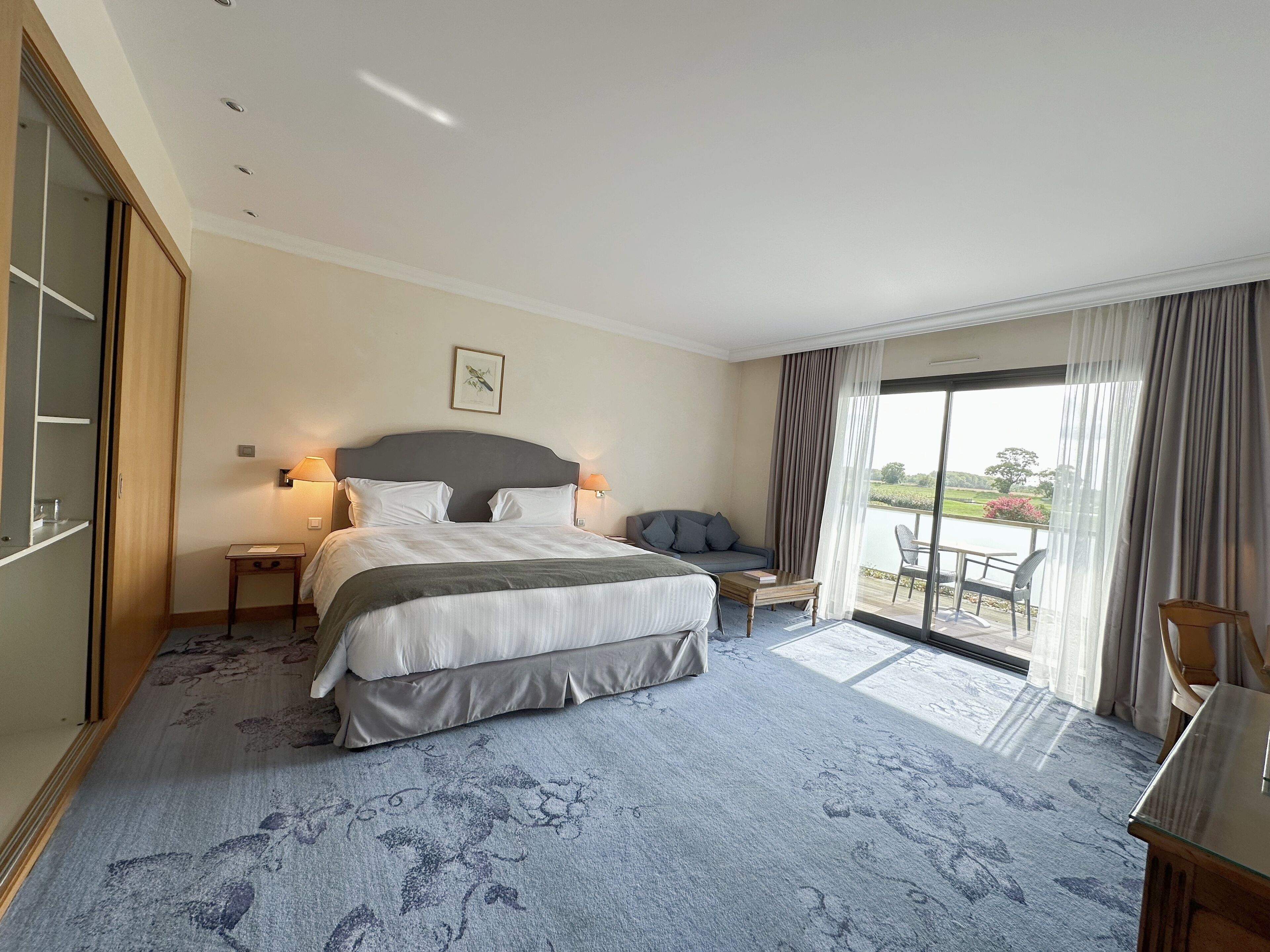 A hotel room with a large bed, a sofa, a small table, and a view of the outdoors.