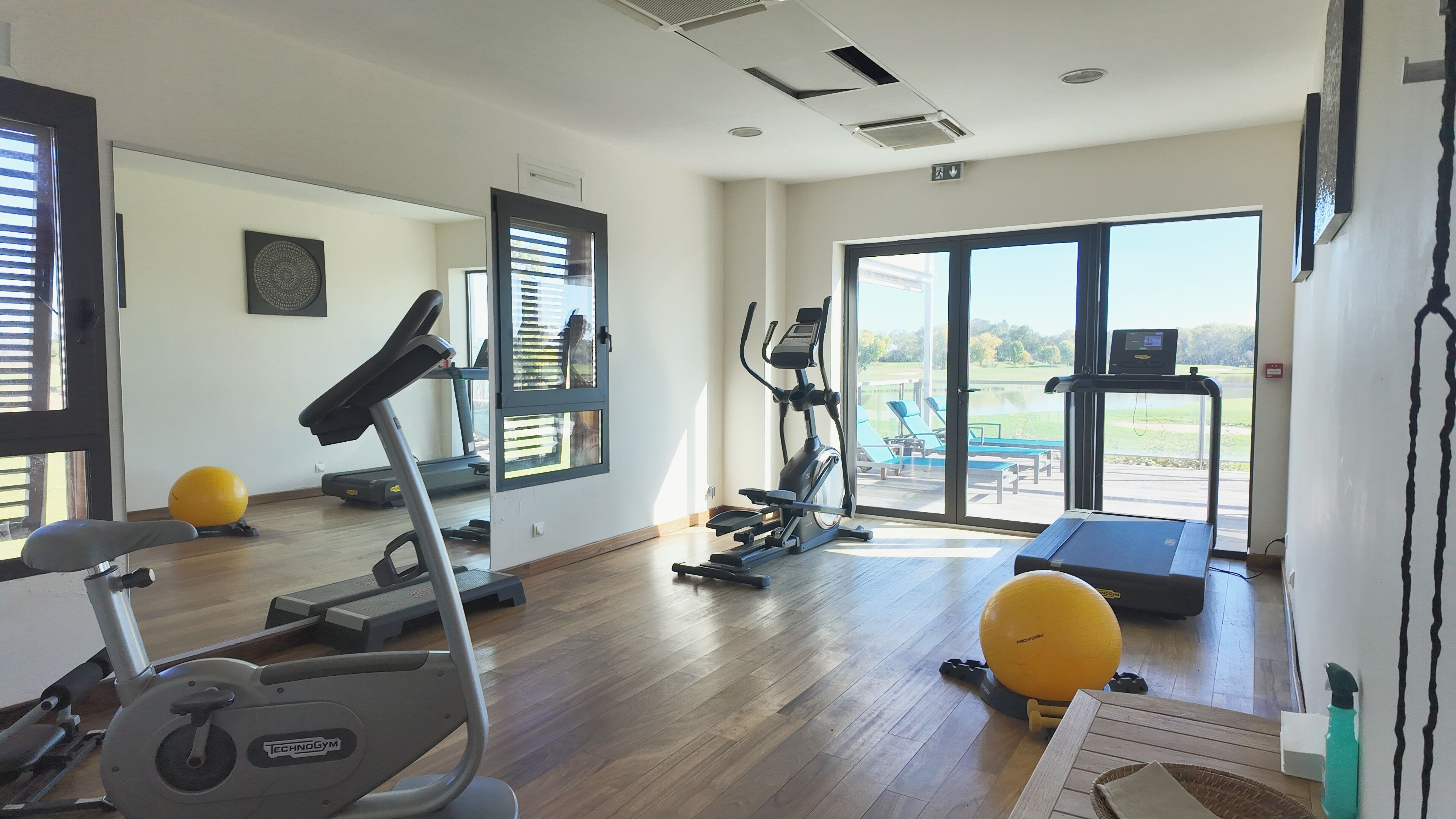 Une salle de sport équipée de matériel d’exercice, dotée d’une grande fenêtre et offrant une vue sur une piscine et un espace extérieur.