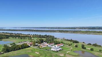 An aerial view of a golf course with a clubhouse, ponds, and a large body of water in the background.