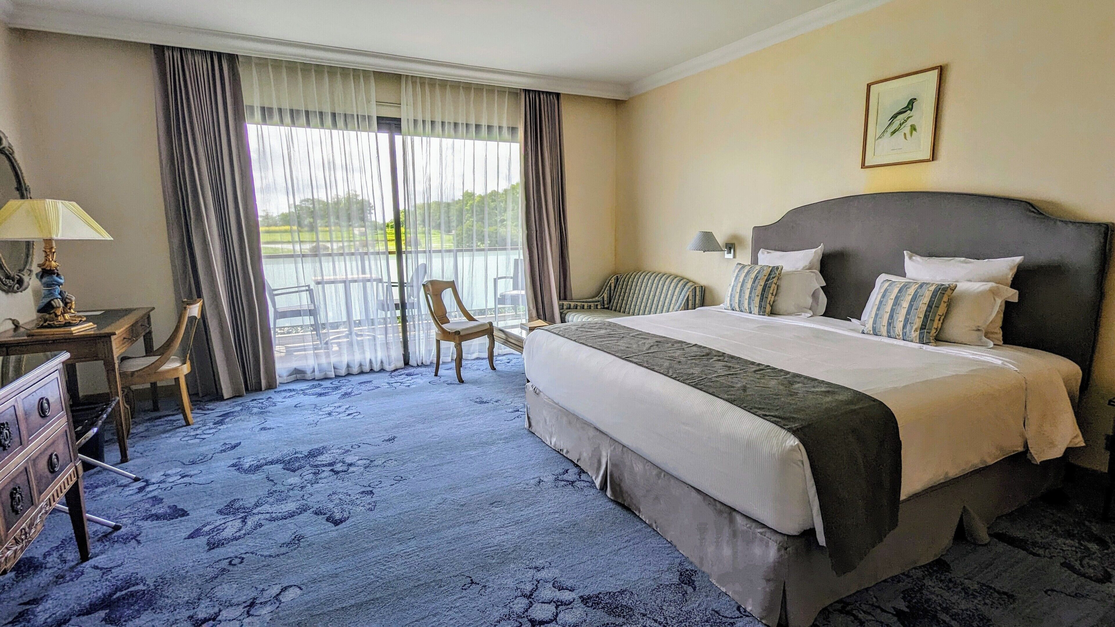Une chambre d’hôtel avec un grand lit, un bureau avec une lampe, une chaise et une vue sur la verdure par la fenêtre.