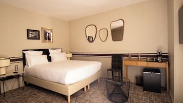 A hotel room with a bed, bedside table, lamp, desk, chair, trash bin, and a refrigerator.