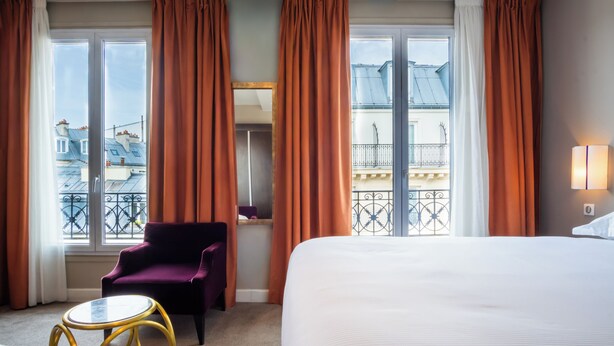 A hotel room with a bed, a chair, a table, and large windows with curtains.