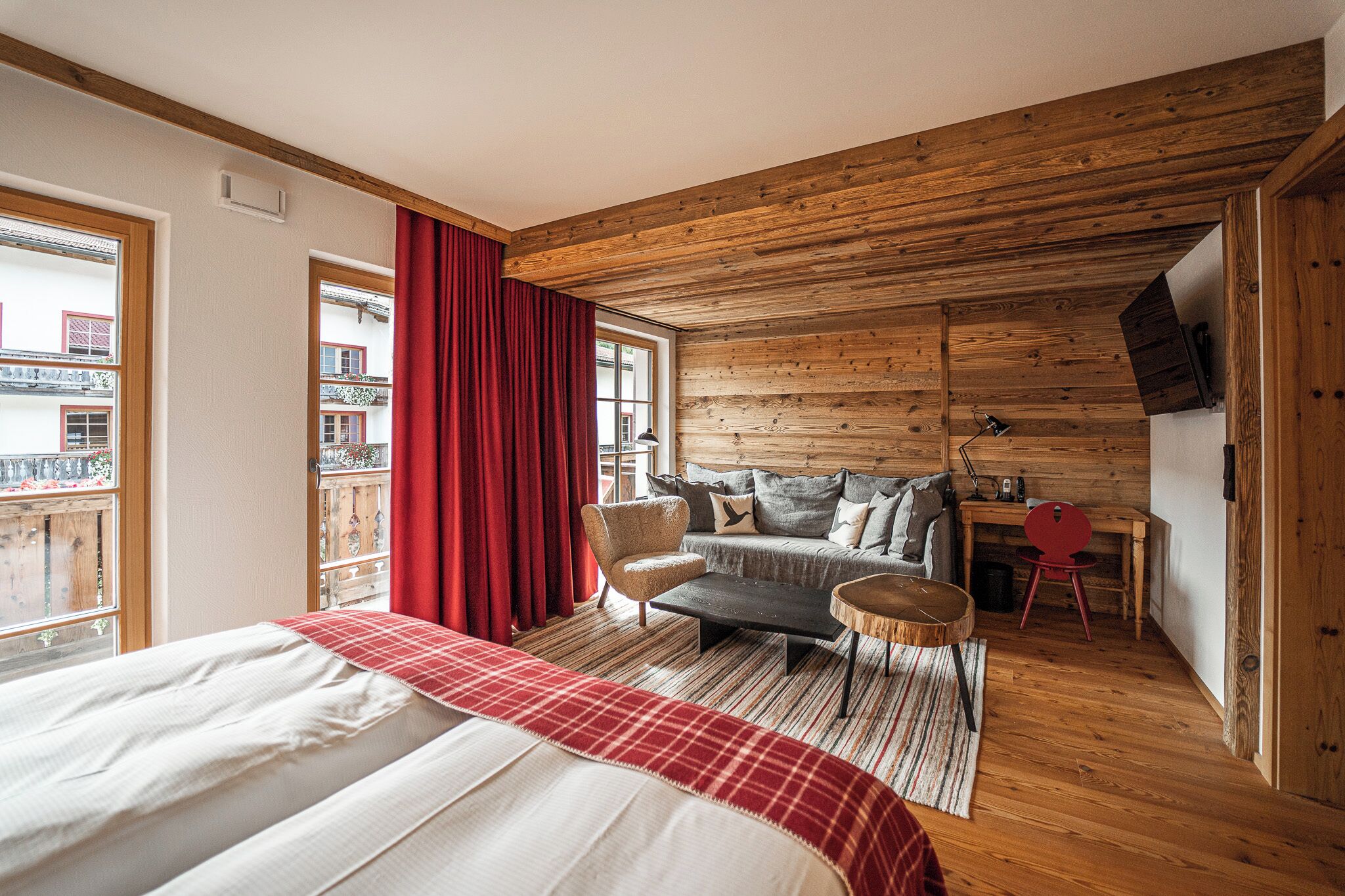 Une chambre à coucher avec un lit, un canapé, une petite table et une chaise. Il y a une fenêtre avec des rideaux, un plafond en bois et une vue sur les bâtiments à l’extérieur.