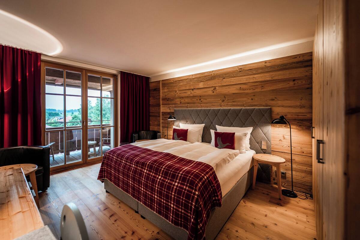 A bedroom with a large bed, a wooden headboard, a nightstand, a desk, and a view through a sliding door.