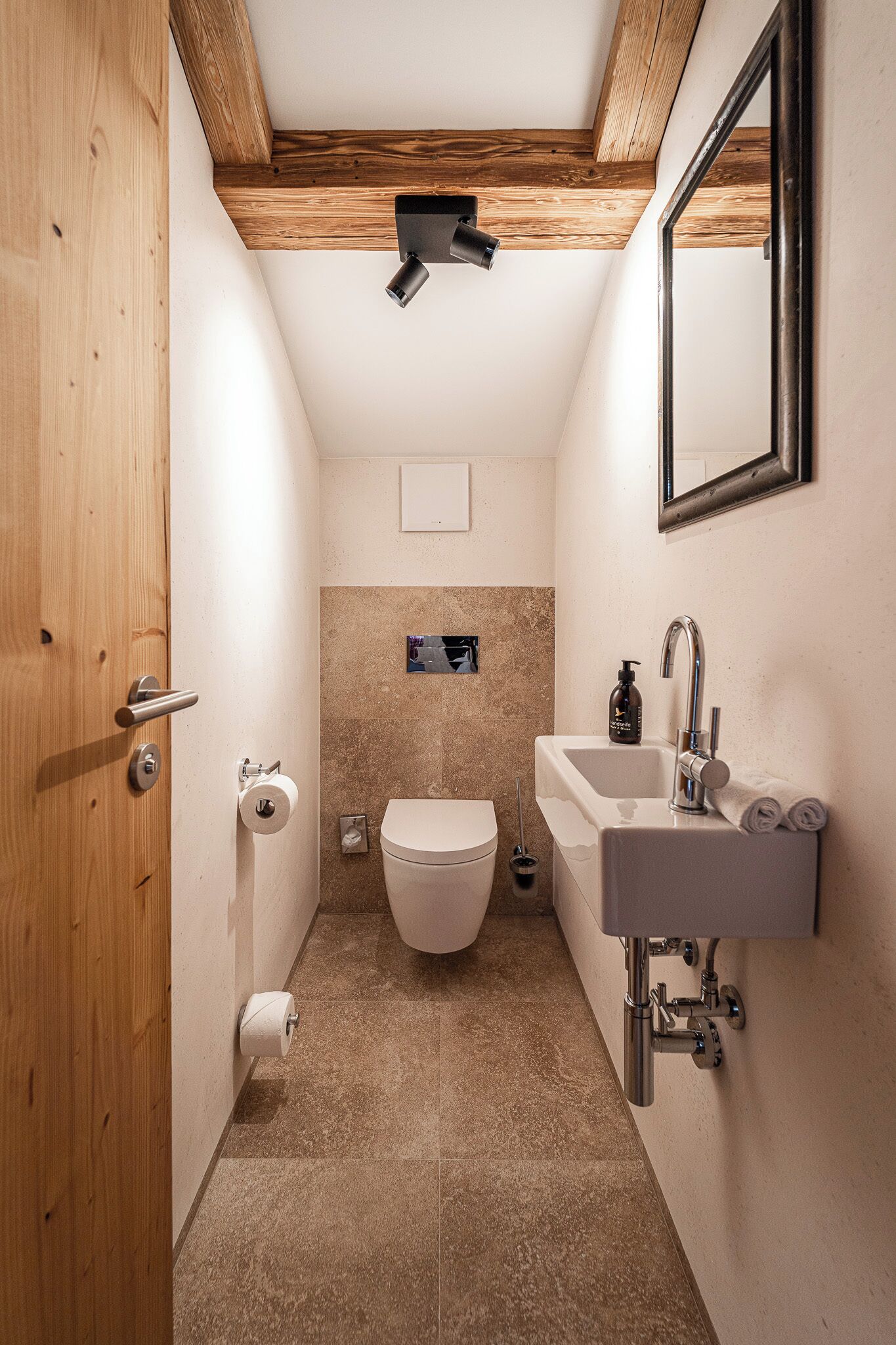 Un baño estrecho con puerta de madera, inodoro, lavabo con espejo y una lámpara en el techo.