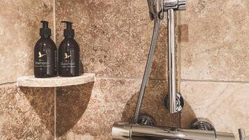A shower area with a chrome faucet and a handheld showerhead. Two bottles of shampoo and a bottle of dish soap are placed on a shelf.
