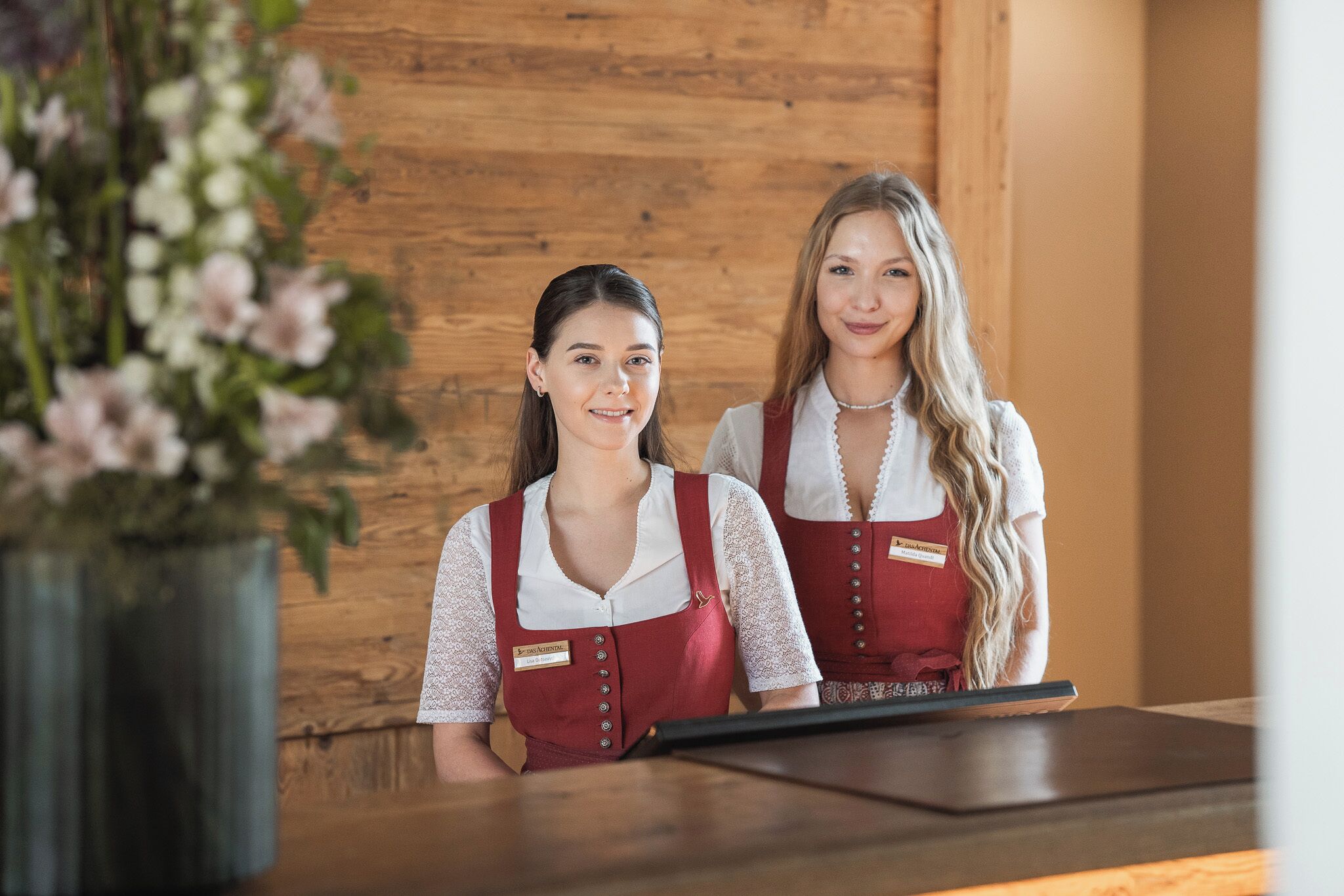 Two receptionists in red aprons with name tags, standing behind a wooden reception desk.