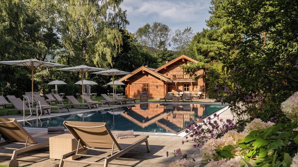 A pool area with lounge chairs, umbrellas, and a wooden cabin. - Resort Das Achental (Grassau)