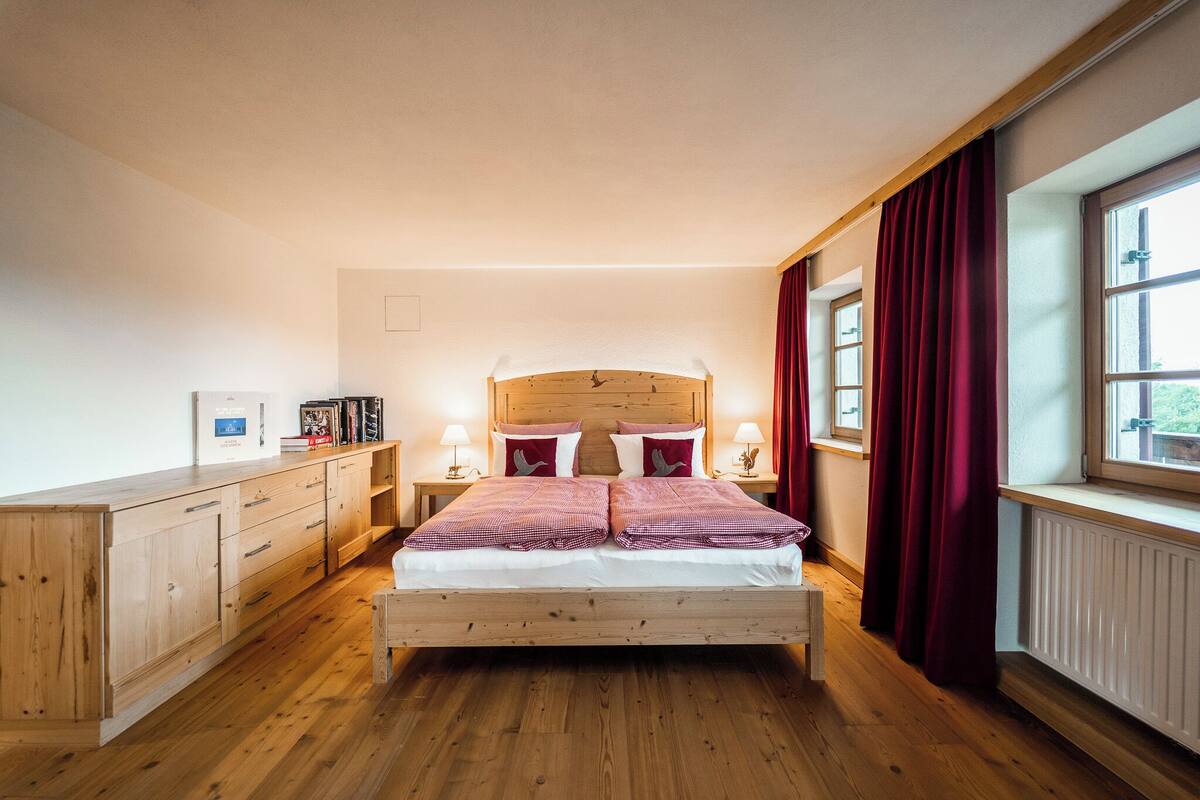 A bedroom with a wooden bed, two bedside tables, a nightstand, a window with curtains, and a wooden wardrobe.