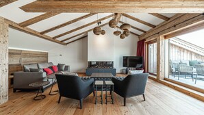 A spacious living area with a fireplace, wooden beams, and a large window. - Resort Das Achental (Grassau)