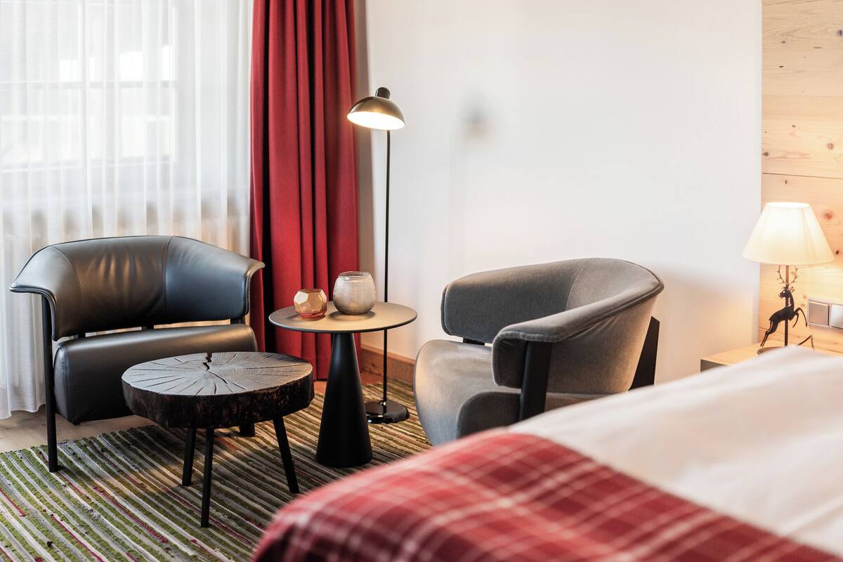 A hotel room with a bed, two chairs, a small round table, a lamp, and a window with curtains.