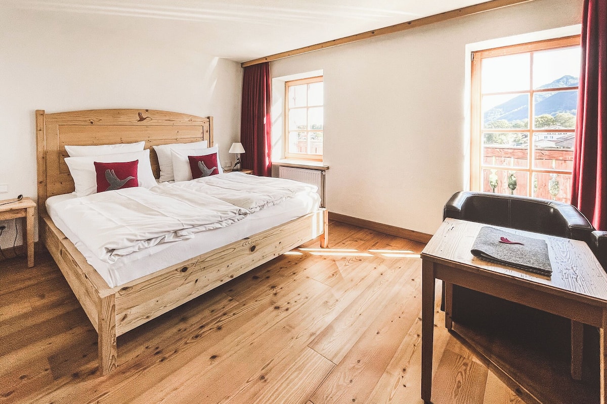 A wooden bed with white bedding, a wooden nightstand, a wooden desk, and a window with a view of buildings and mountains.