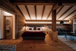 A bedroom with a wooden ceiling, a bed with pillows, a nightstand, a lamp, and a sofa. - Resort Das Achental (Grassau)