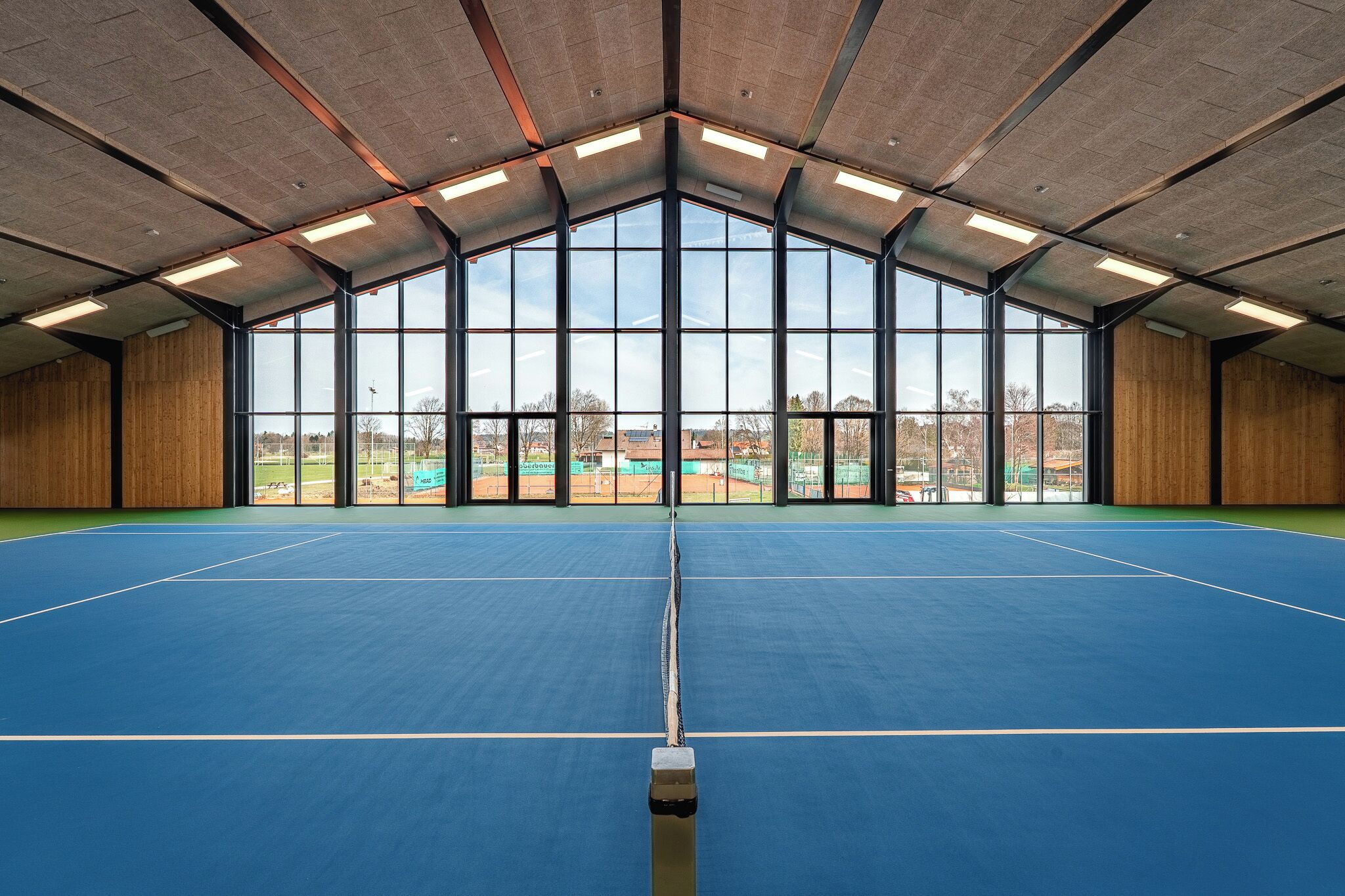 Un court de tennis intérieur doté d’un plafond haut, de grandes fenêtres et offrant une vue sur l’extérieur.