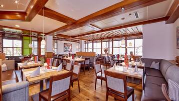 Ein Restaurant mit Holztischen und -stühlen, großen Fenstern und einer hohen Decke mit freiliegenden Balken.