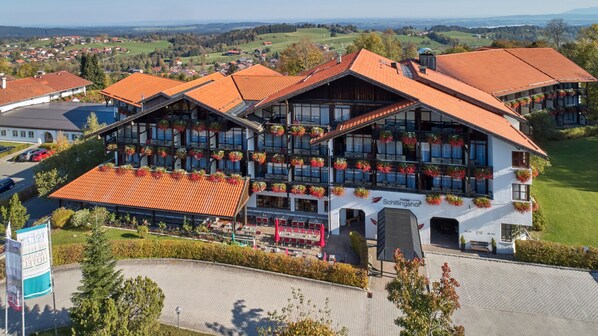 A hotel with a traditional Alpine architectural style, featuring a large balcony with flower boxes, a covered outdoor seating area, and a view of the surrounding countryside.