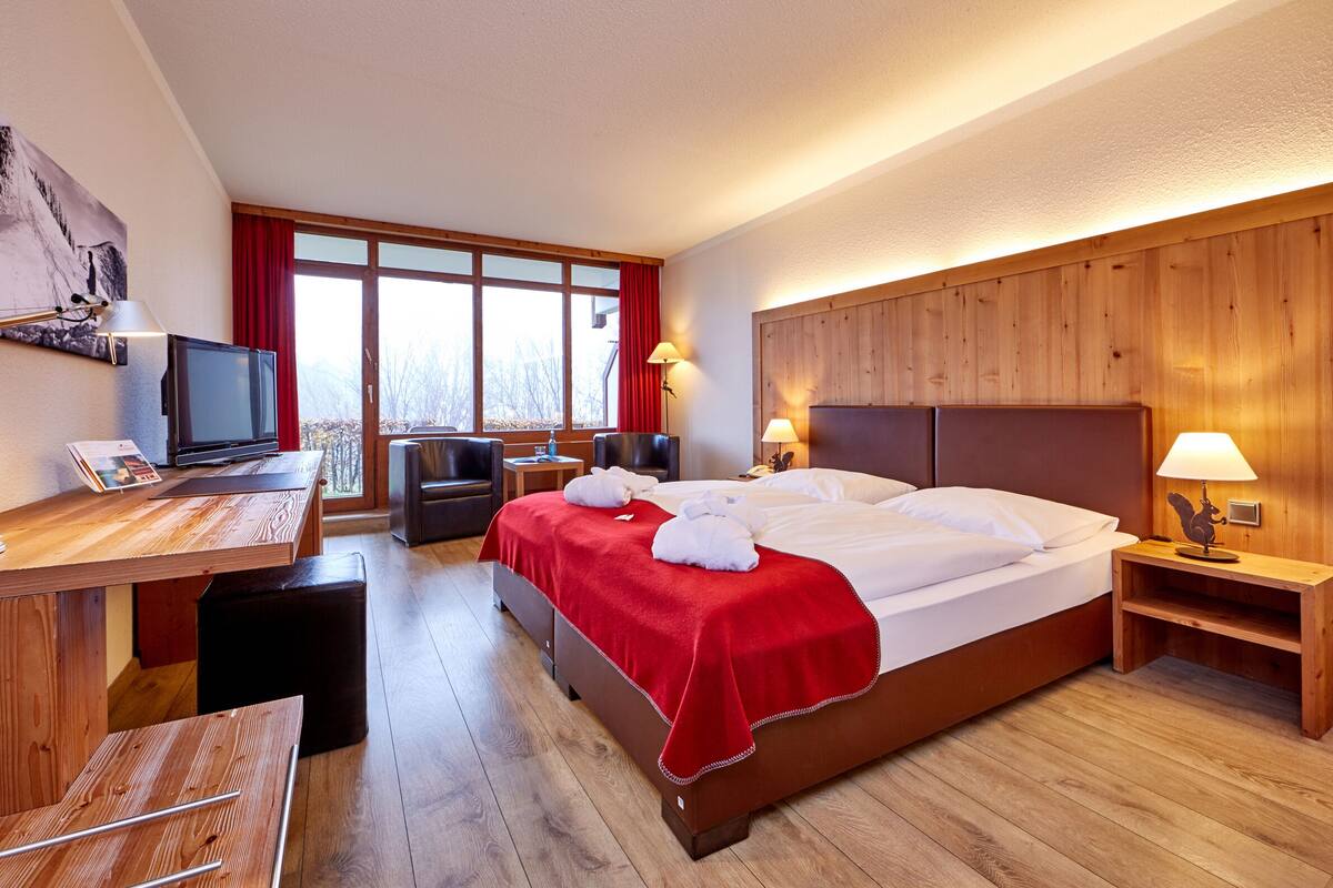 Een hotelkamer met een houten hoofdeinde, een bureau met een computer, een bed met witte lakens en een rode plaid.