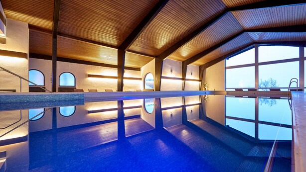 Una piscina interior moderna con vigas de madera, ventanas amplias y una vista despejada del cielo.