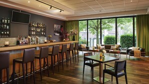 Zona de bar con una barra alargada y varios taburetes, un área de estar con una mesa y sillas, y grandes ventanales con vistas a edificios y zonas verdes.