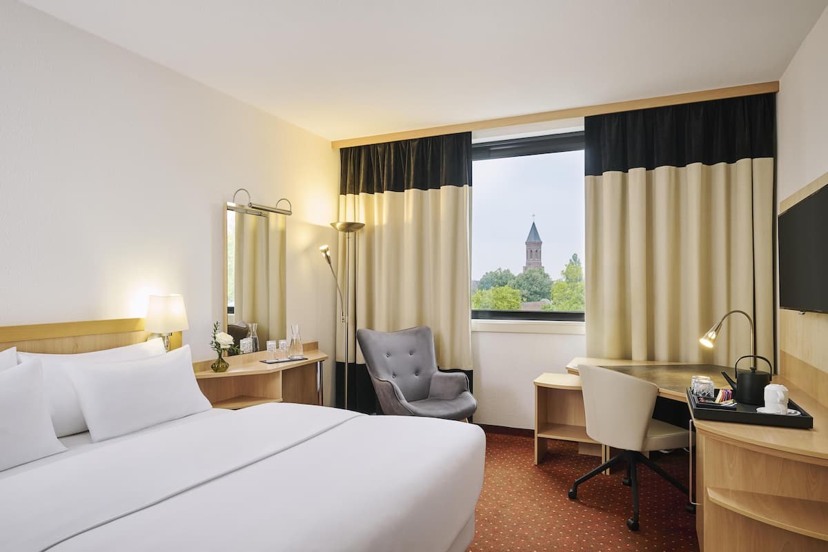 A hotel room with a bed, desk, chair, and a view of a building through the window.