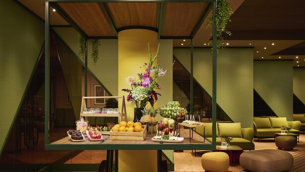 Un interior moderno con una mesa central que exhibe frutas y postres, asientos tapizados en verde y un suelo con estampado geométrico.