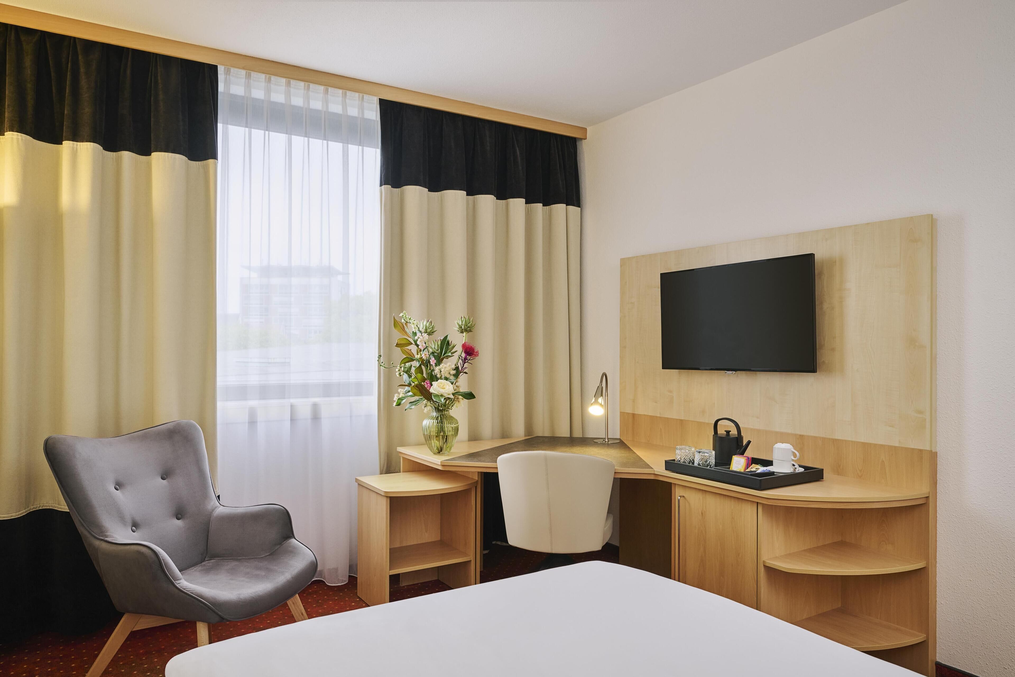 A hotel room with a bed, a desk with a TV, a chair, a vase with flowers, and a window with curtains.