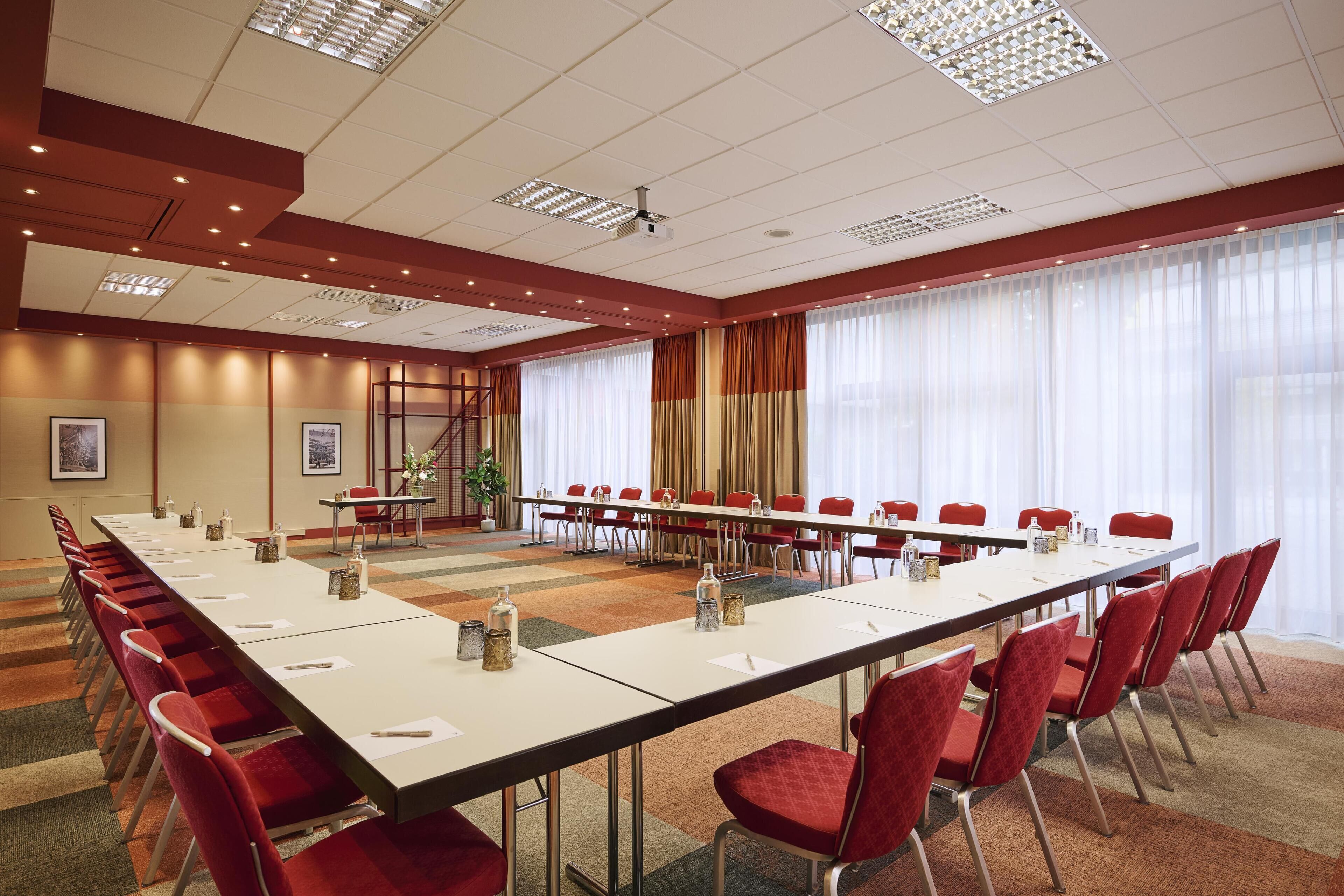 Un salón de conferencias con una larga mesa, sillas rojas y una pantalla para proyector.