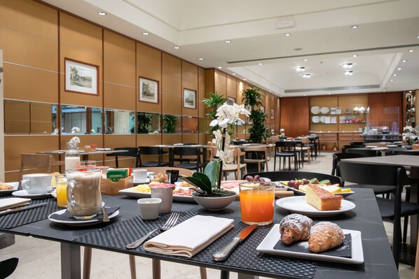 A hotel dining area with tables set for breakfast, featuring a variety of dishes, drinks, and a decorative orchid plant.