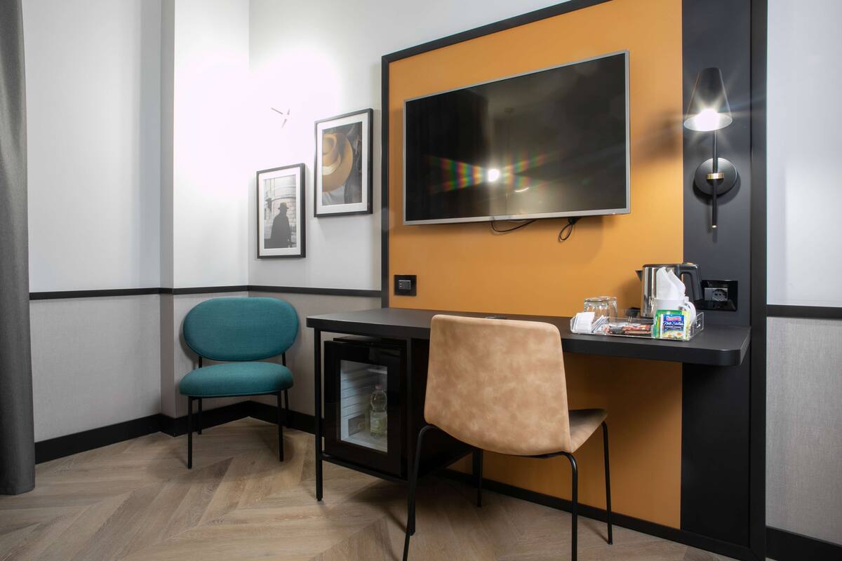 A hotel room with a TV mounted on the wall, a small table with a chair, and a desk with a chair.