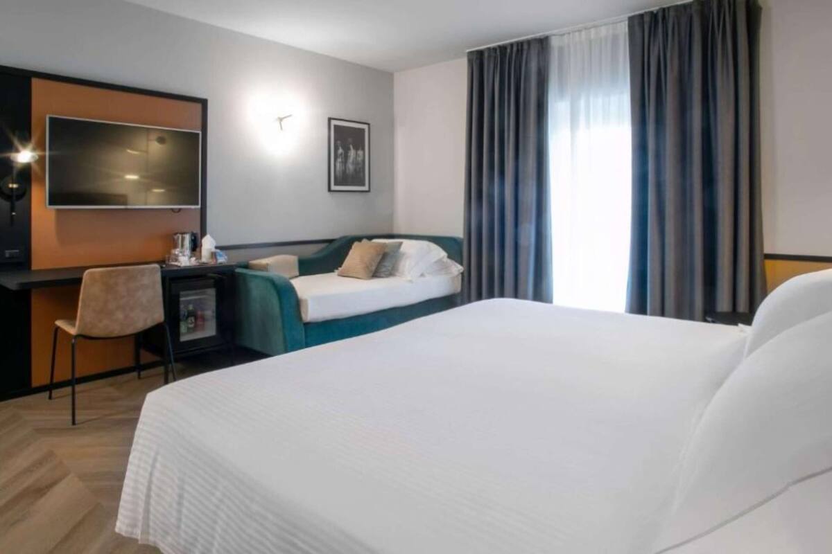 A hotel room with a large bed, a desk with a chair, a flat-screen TV, and a window with curtains.