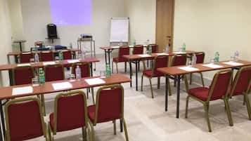 Ein Konferenzraum mit mehreren Tischen und Stühlen, ausgestattet mit Wasserflaschen und Unterlagen, einer Projektionsleinwand und einer Whiteboard-Tafel.