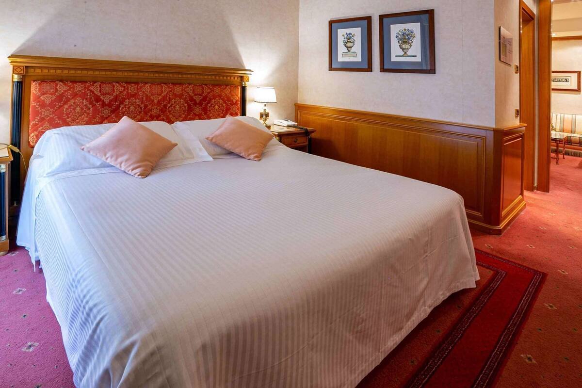 A hotel room with a large bed, a wooden headboard, a nightstand, a lamp, and two framed pictures on the wall.