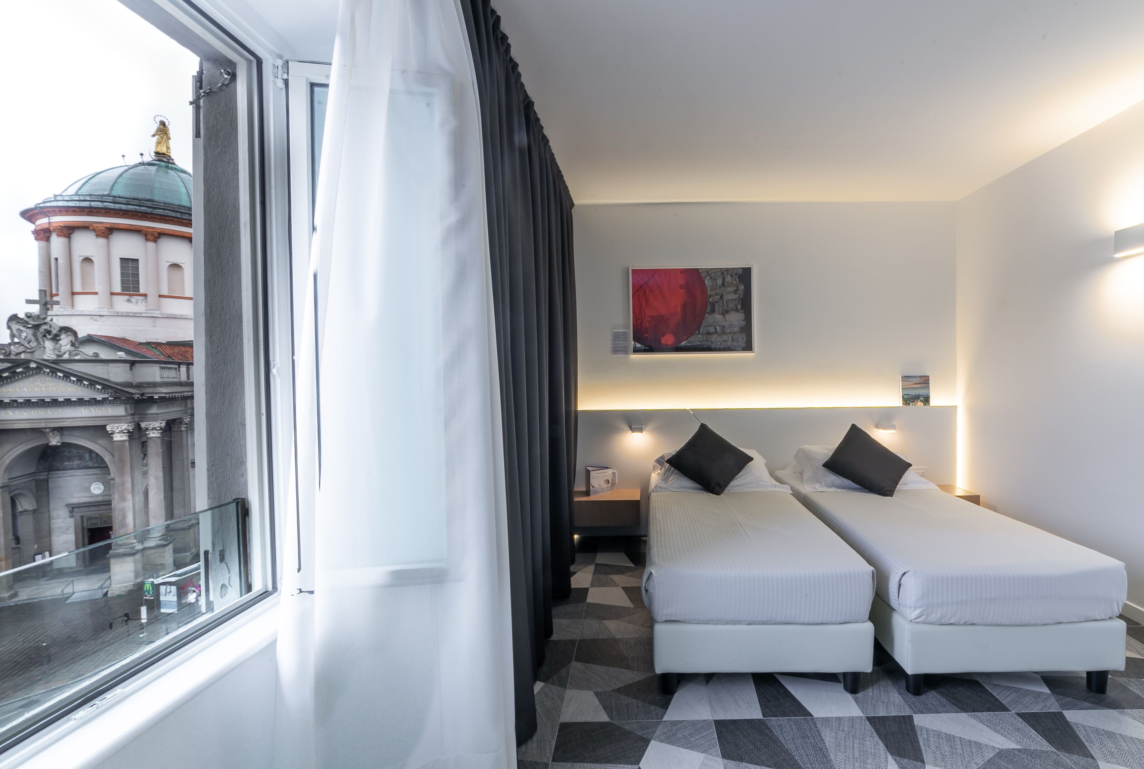 Hotelkamer met twee enkele bedden, een groot raam met gordijnen, een receptiebalie en uitzicht op een historisch gebouw.