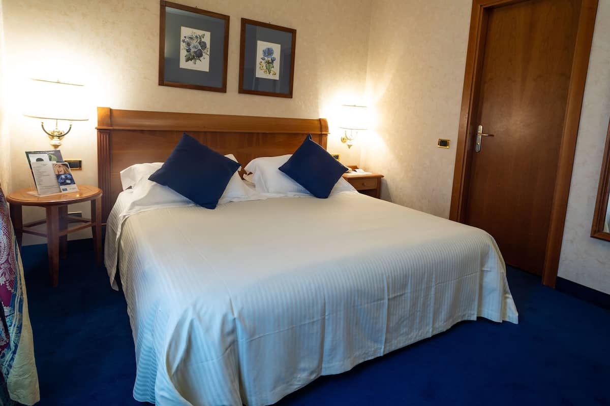 A hotel room with a large bed, two bedside tables, a nightstand, a door, and framed pictures on the wall.