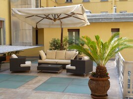 Zona salotto esterna con divano, sedie e tavolo, circondata da piante in vaso e un ombrellone.