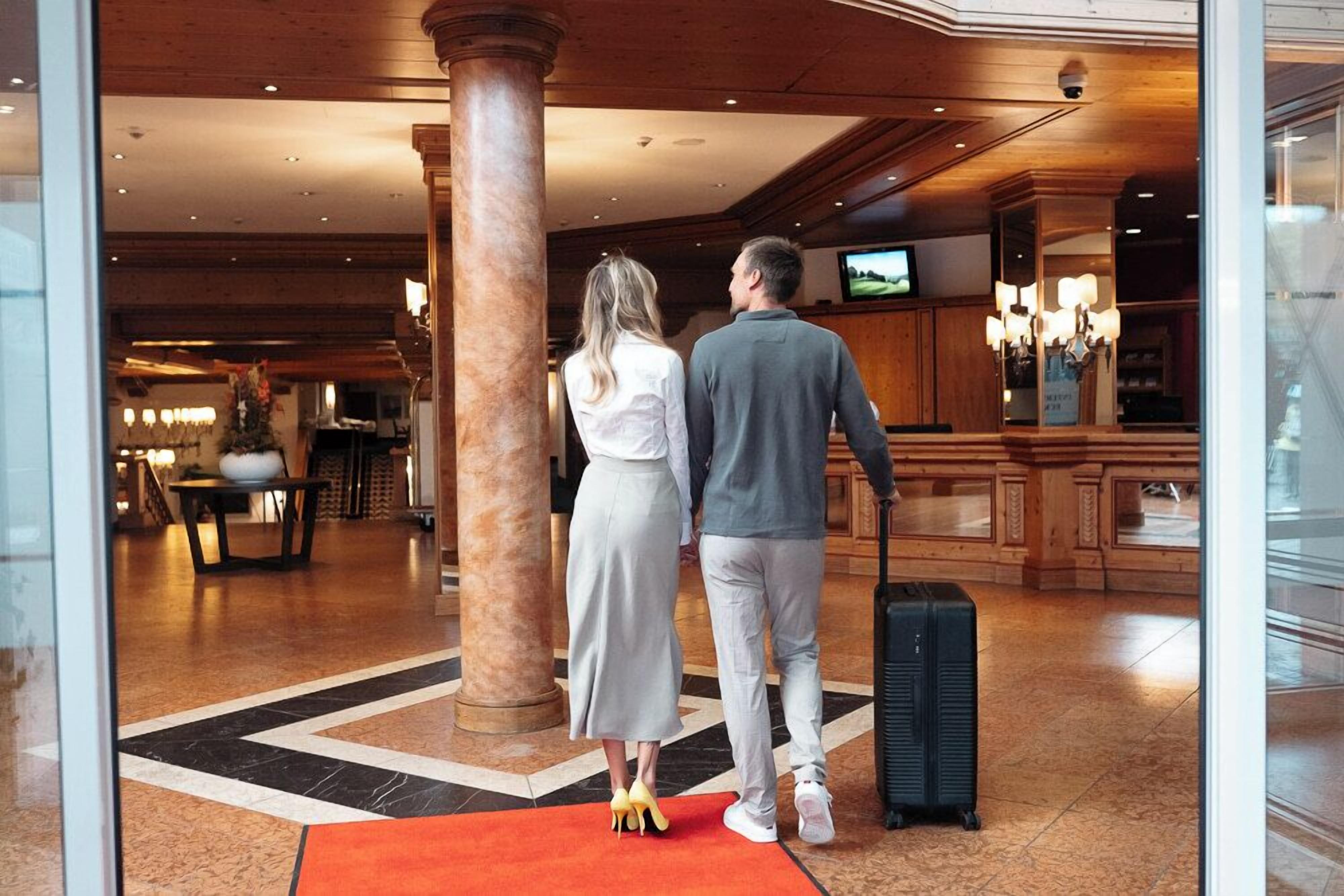 Un couple se tient dans un hall d’entrée, devant un tapis rouge, une grande colonne et un téléviseur accroché au mur.