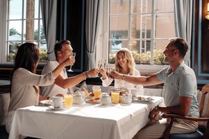A group of people toasting at a table with breakfast items, including orange juice and pastries, in a room with large windows and curtains.