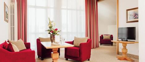 A hotel room with a dining table, red sofas, a TV, and a large window with curtains.