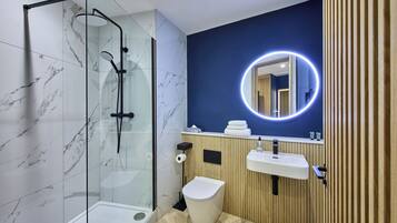 Un baño moderno con ducha envuelta en vidrio, inodoro blanco, tocador de madera con lavamanos y espejo con luz circular.